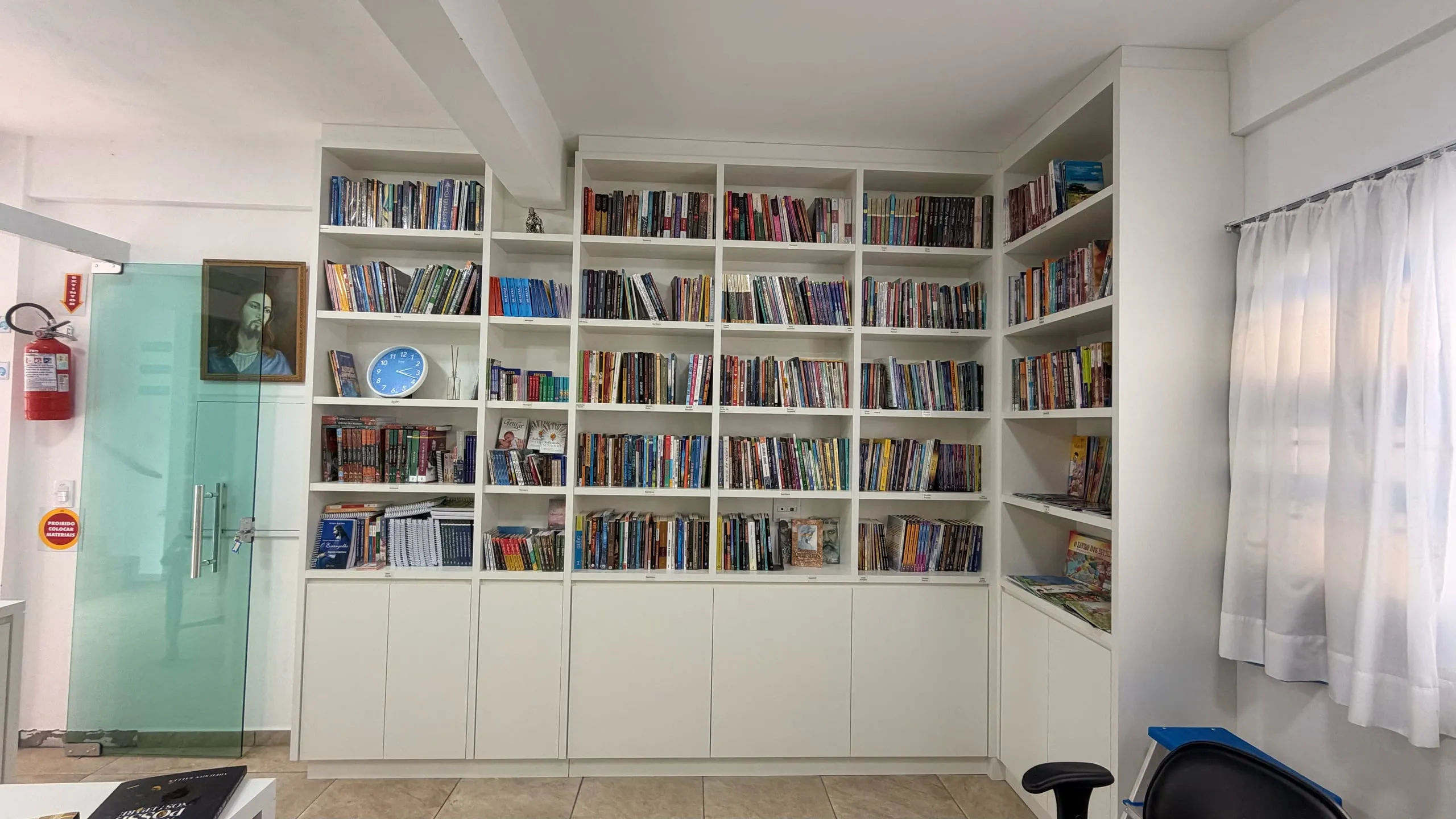 A livraria do CECAL: estantes brancas do chão ao teto cheias de livros, uma porta de vidro à esquerda e a luz da janela entrando pela cortina.