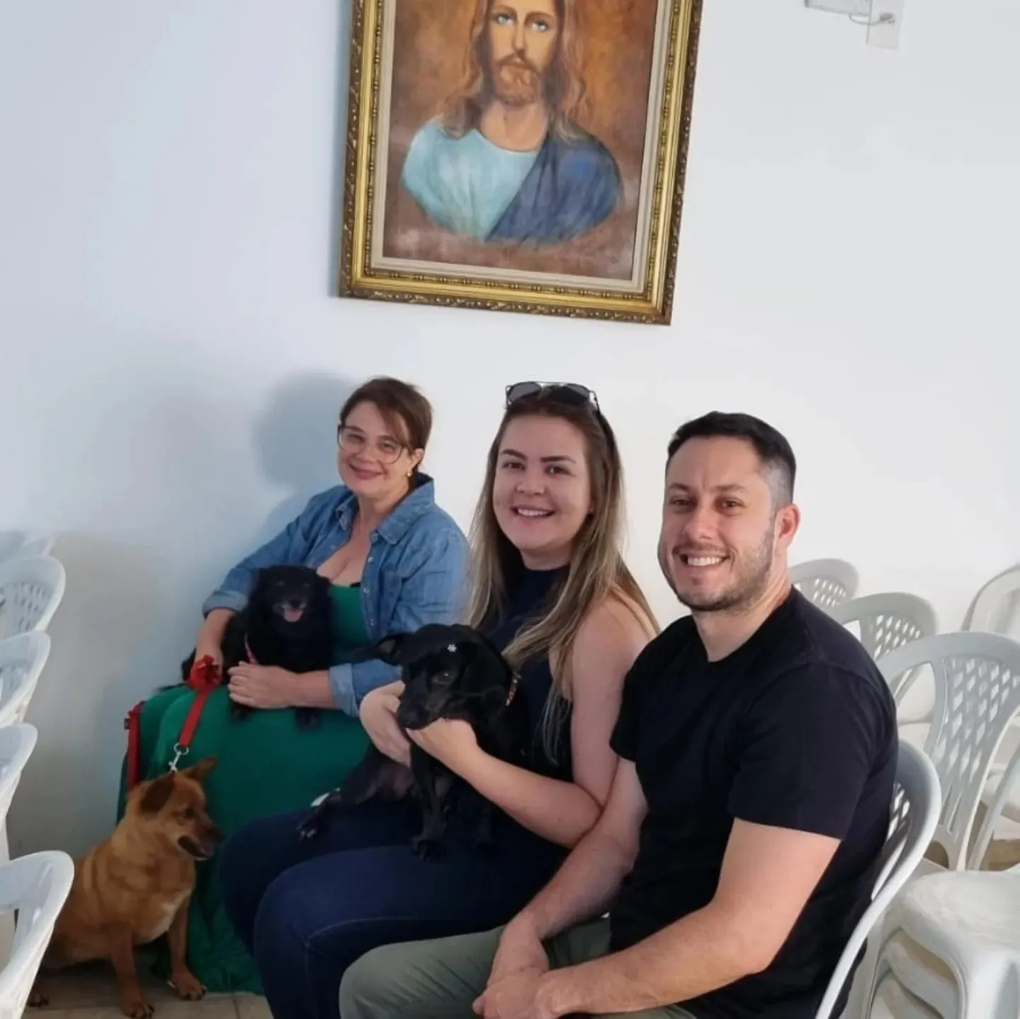 Três pessoas sentadas no salão do CECAL com seus cães, sob o quadro de Jesus.