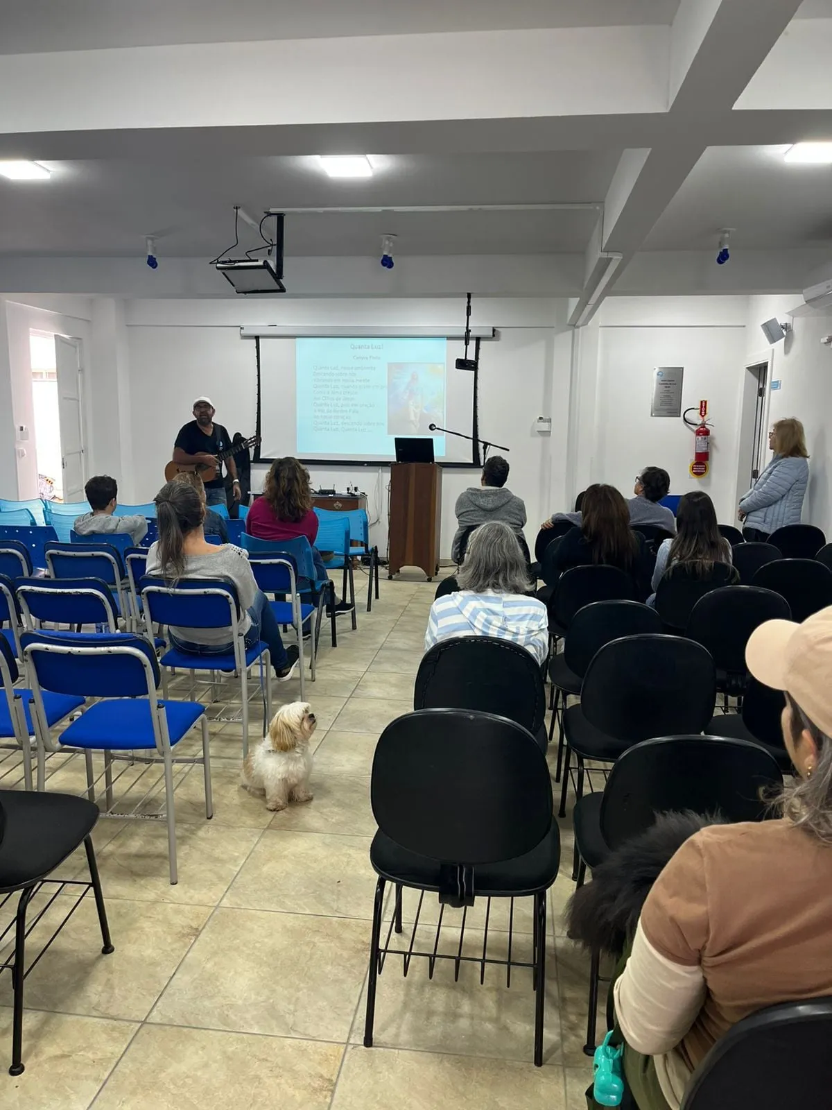 Salão do CECAL durante uma palestra: fileiras de cadeiras pretas e azuis ocupadas, o palestrante à frente ao lado da tela projetada, e um cachorro pequeno deitado entre as fileiras.