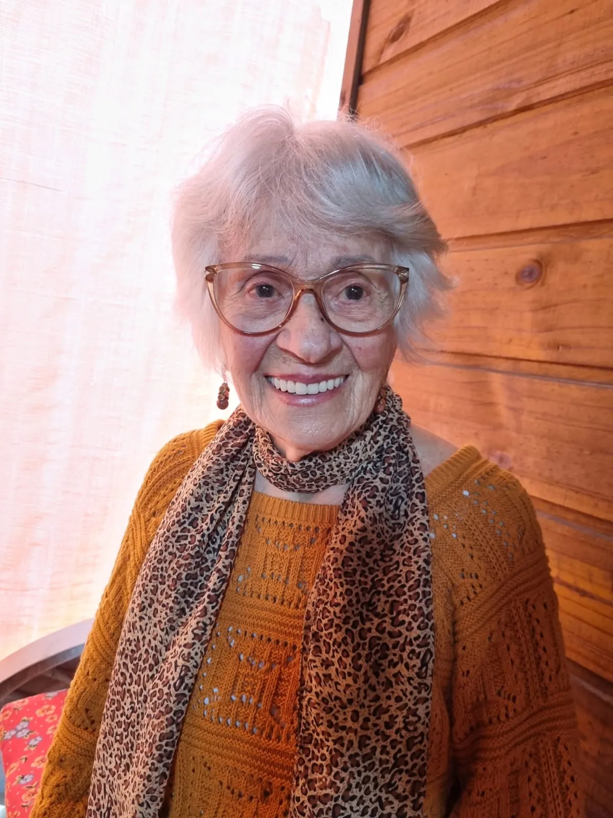Retrato de uma senhora de cabelos brancos e óculos, com um lenço estampado no pescoço, sorrindo.