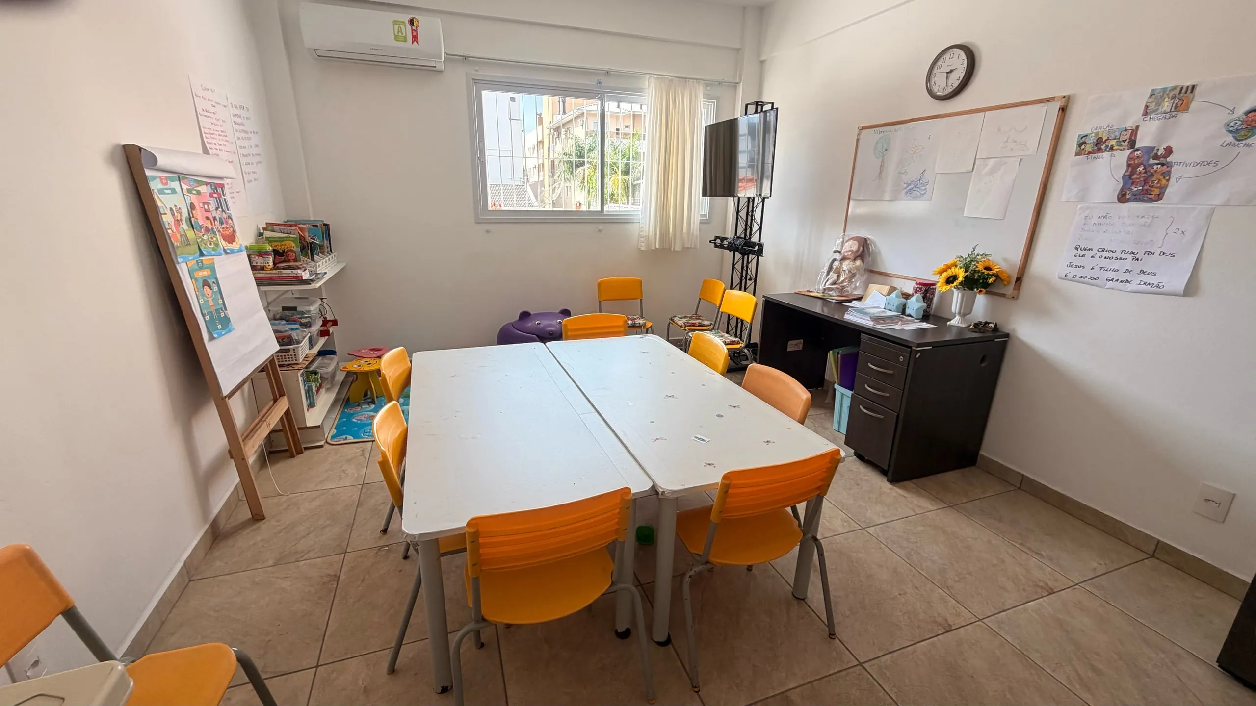 A sala da evangelização arrumada: mesas brancas, cadeiras laranja e a lousa na parede.