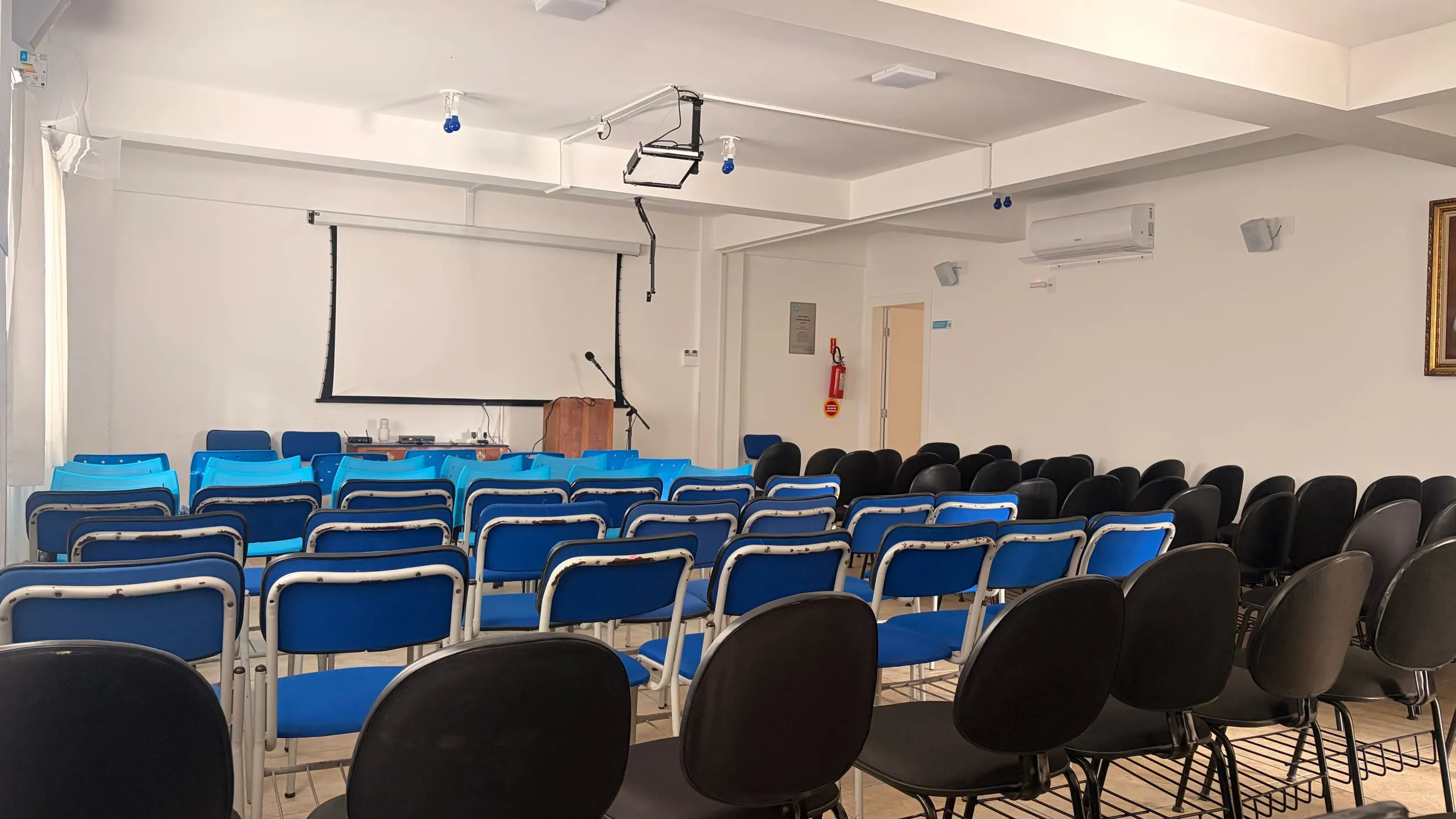 O salão do CECAL vazio, visto do fundo: fileiras de cadeiras pretas e azuis voltadas para a tela e o púlpito, ao centro.