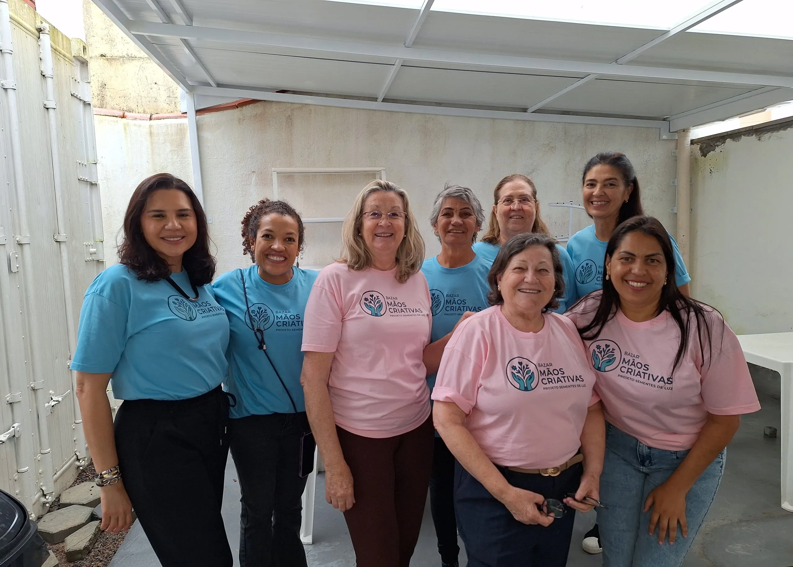 Oito voluntárias do Sementes de Luz sorrindo no pátio da casa, com as camisetas do Bazar Mãos Criativas.